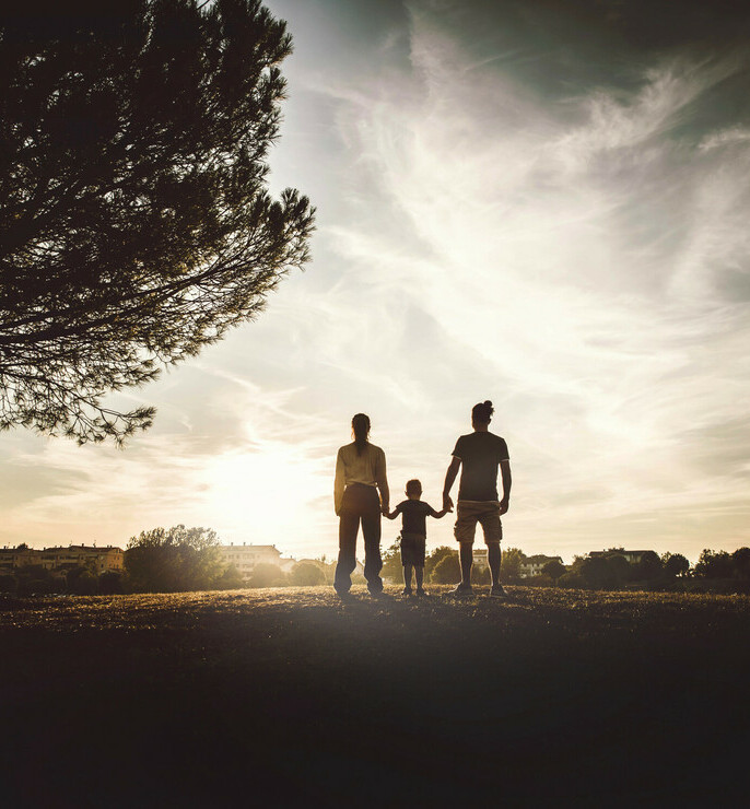 Silhouettes of two parents and one small child standing in the distance by a tree watching the sunset